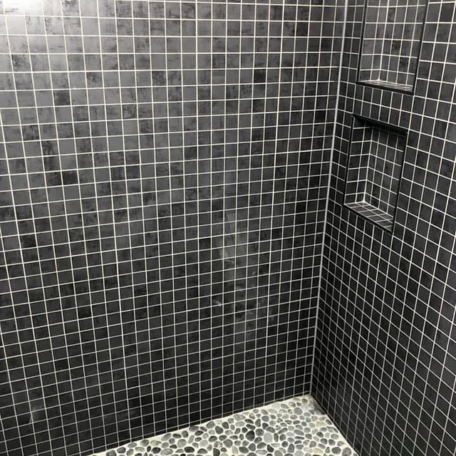 Dusjsone med mørke mosaikkfliser og gulv av rullestein, med praktiske hyller i vegg.